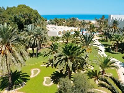 Grüner Garten mit Palmen, blauer Himmel und Meer im Hintergrund, weiße Wege und Gebäude.
