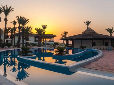 Une piscine d'hôtel calme au coucher du soleil avec des palmiers et des pavillons.
