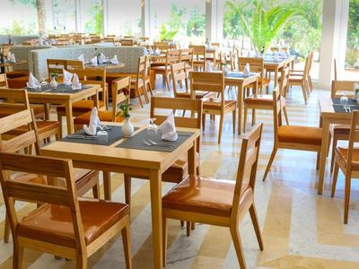 Helles Restaurant mit Holztischen und Stühlen, eingedeckt mit Servietten und Gläsern, viele Fenster mit Pflanzenblick.