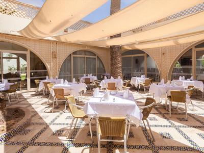 Freiluftrestaurant mit rundem Holztischen, weißen Tischdecken und Korbstühlen unter hellen Sonnensegeln.