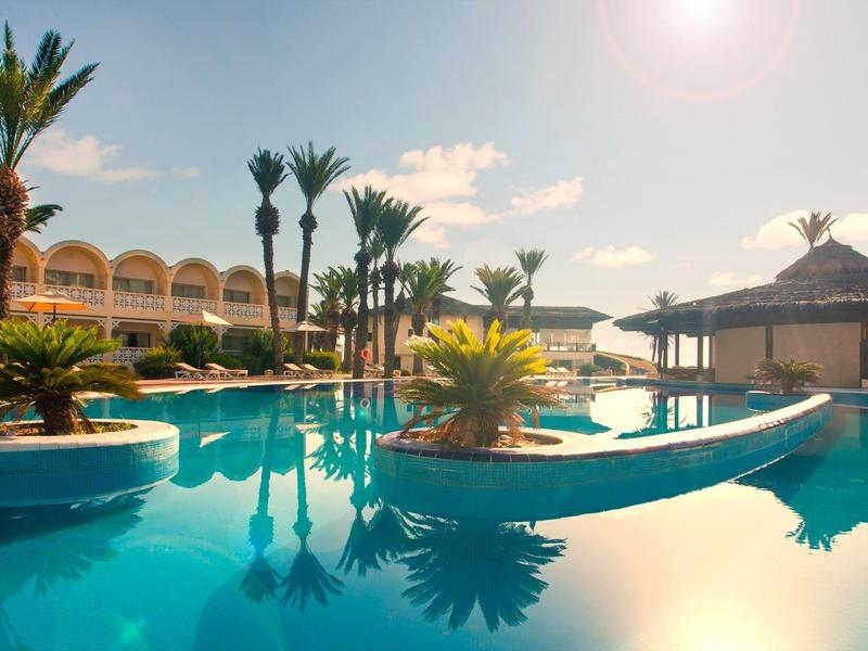 Großer Pool mit Palmen, Sonnendach und mehrstöckigem Hotel bei sonnigem Himmel.