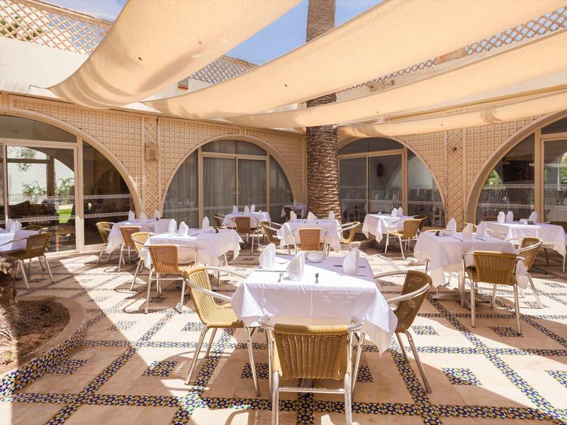 Freiluftrestaurant mit rundem Holztischen, weißen Tischdecken und Korbstühlen unter hellen Sonnensegeln.