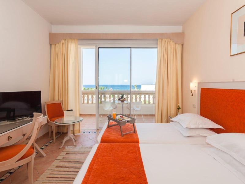 Hell eingerichtetes Hotelzimmer mit zwei Betten, orangefarbenen Akzenten und Meeresblick durch Fenster.