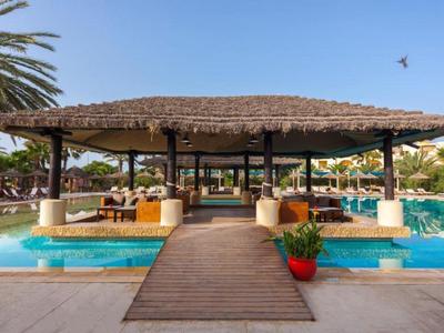 Offenes Poolrestaurant mit Holzsteg, Palmen, blauen Wasserbecken und roten Pflanzenkübeln.