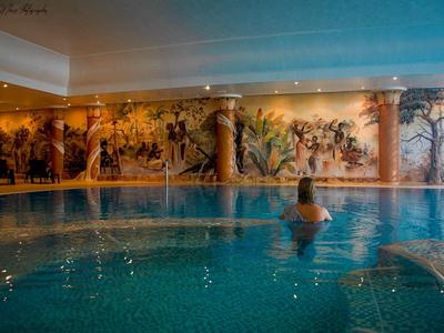 Vnitřní bazén s nástěnnou malbou a osobou relaxující ve vodě pod stropním osvětlením.