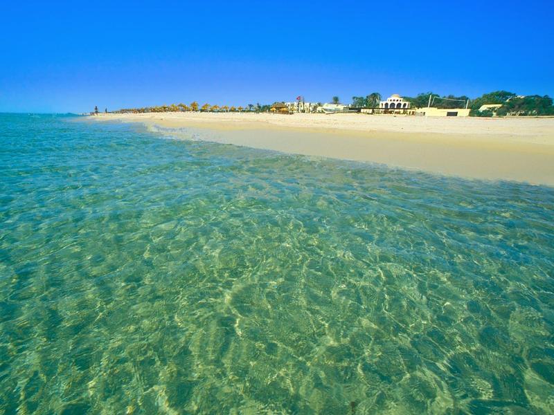 Čirá tyrkysová voda podél písčité pláže pod jasně modrou oblohou.