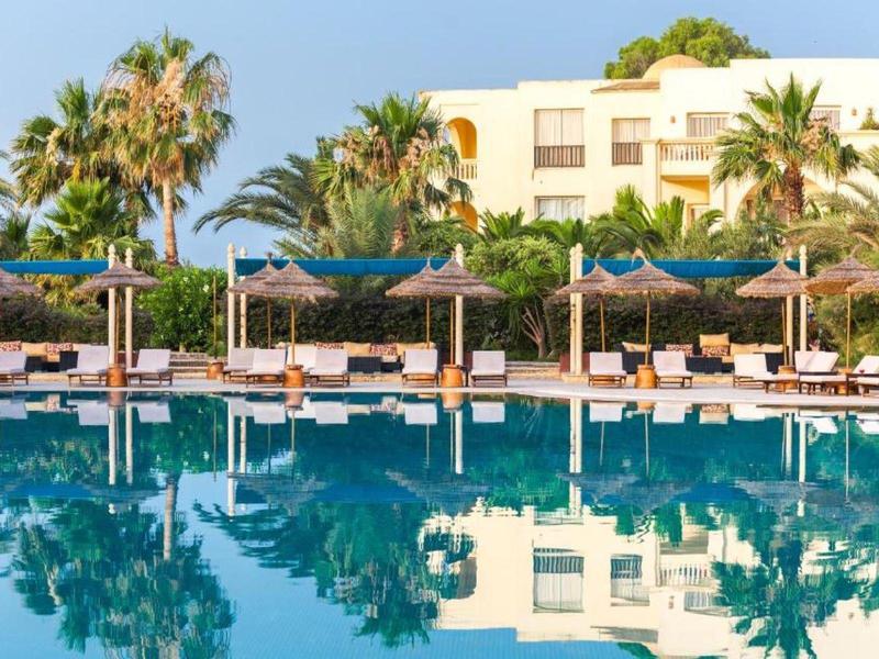 Bazén v rezortu s lehátky, slunečníky, palmami a hotelem v pozadí.