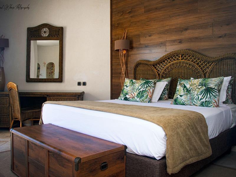 Pohodlný hotelový pokoj s dřevěným čelem postele, tropickými polštáři, velkou postelí a vintage truhlou u nohou.
