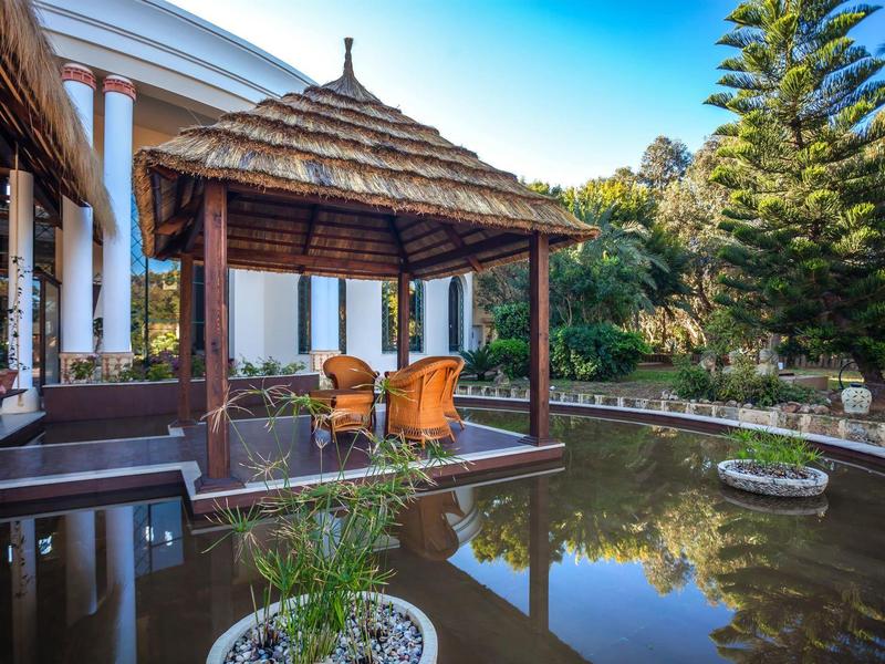 Venkovní altán s židlemi u rybníka obklopeného rostlinami a stromy poblíž bílé budovy.