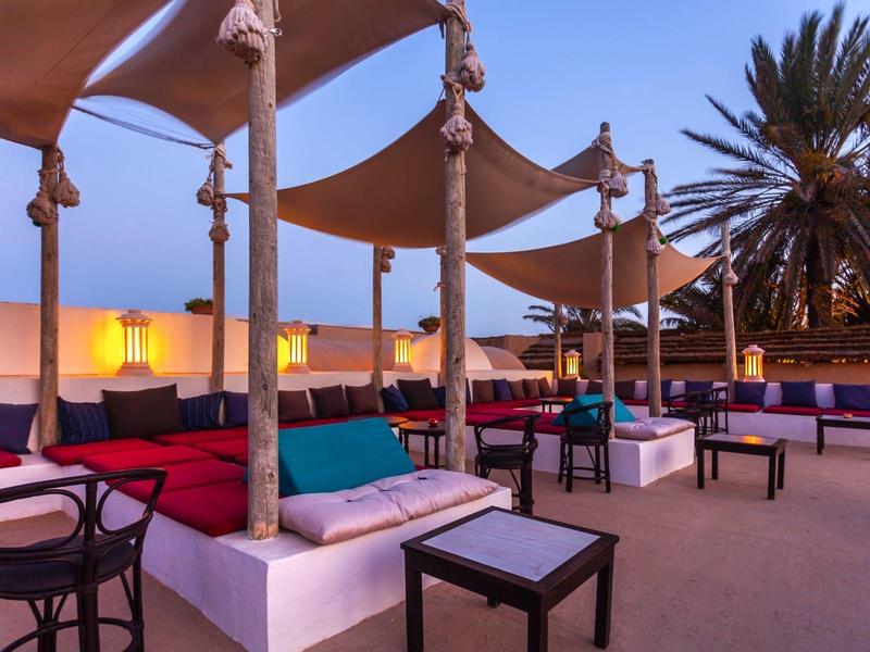 Venkovní lounge s barevnými polštáři, slunečními plachtami a palmami při západu slunce.