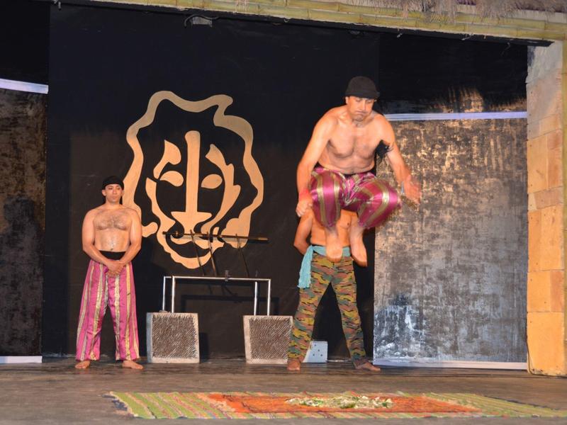 Dva muži v lesklých růžových kalhotách skáčou na jevišti s černým pozadím a emblémem.