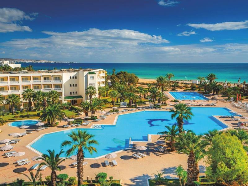 Grande piscine d'hôtel avec transats et palmiers à côté de la plage sous un ciel bleu.