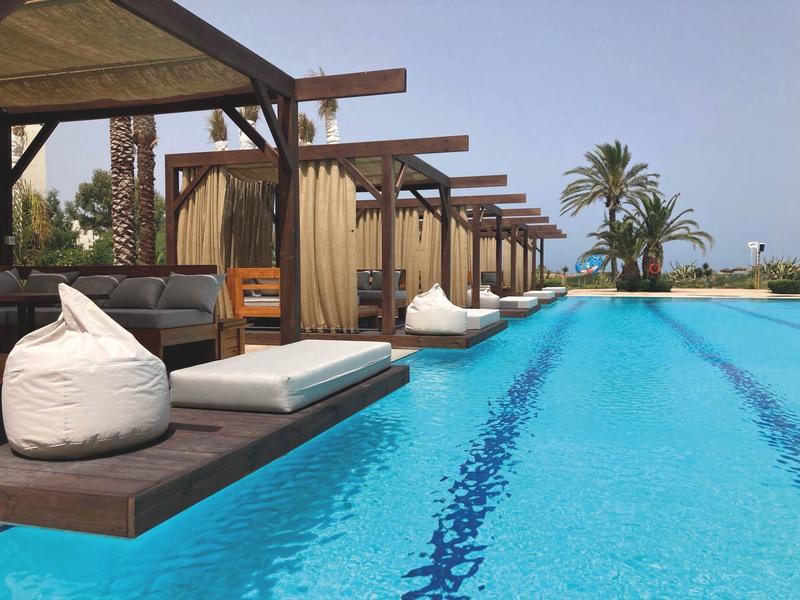 Longue piscine bleue avec transats, poufs et palmiers sous un ciel clair.