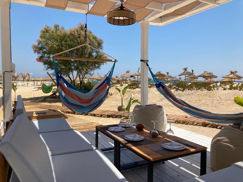 Terrasse de plage avec transats, hamacs et table sous un auvent face à la plage de sable.