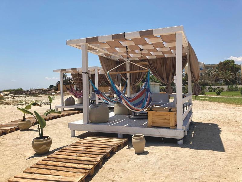 Coin détente confortable sur la plage avec hamacs, plateforme en bois et pare-soleil sous un ciel bleu.