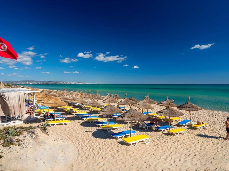 Plage avec transats, parasols, drapeau rouge et ciel bleu clair au bord de la mer.
