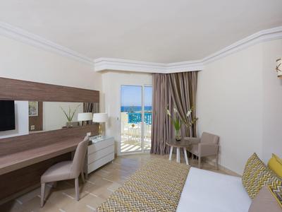 Camera d'hotel moderna con scrivania, poltrona, letto e vista mare attraverso una porta aperta.