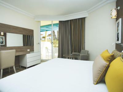 Helles Hotelzimmer mit großem Bett, Schreibtisch, Stuhl und Balkon mit Ausblick.