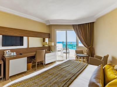 Camera d'hotel luminosa con vista mare, letto, scrivania, sedia e balcone con ringhiera in vetro.