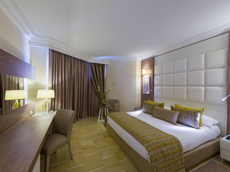 Camera d'hotel moderna con letto matrimoniale, scrivania, tende e illuminazione luminosa.