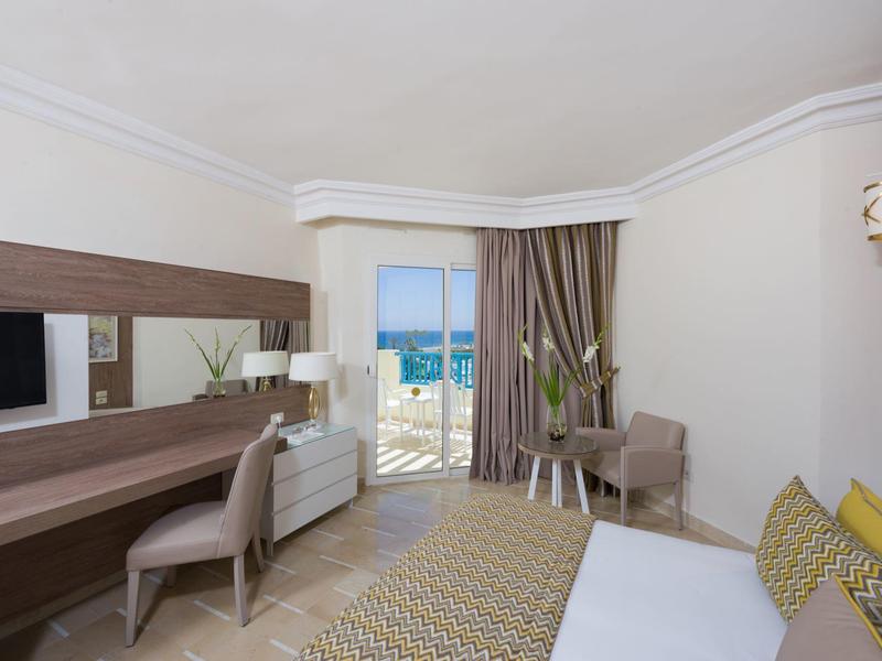 Camera d'hotel moderna con scrivania, poltrona, letto e vista mare attraverso una porta aperta.