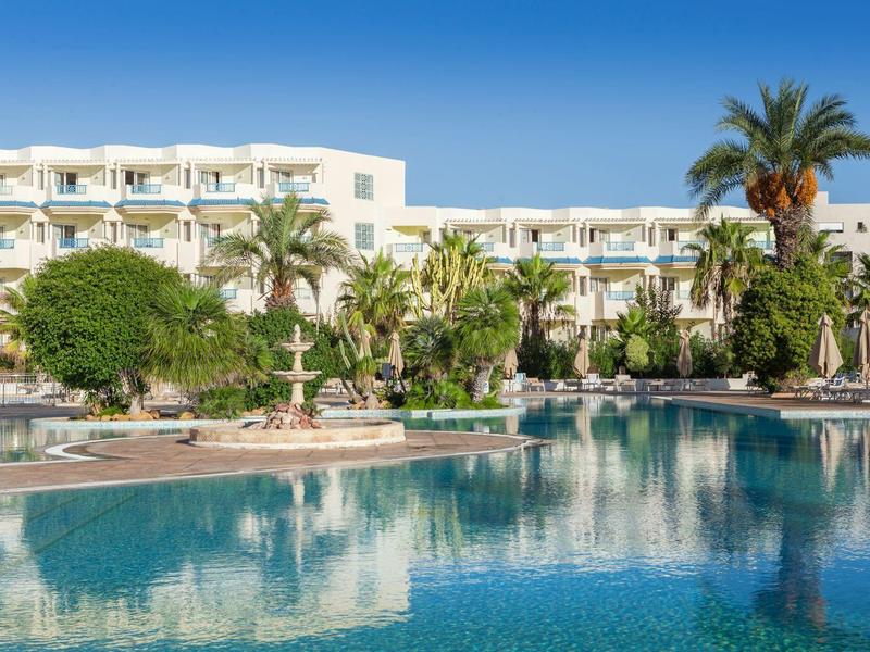 Hotel con piscina, palme e ombrelloni sotto un cielo azzurro.