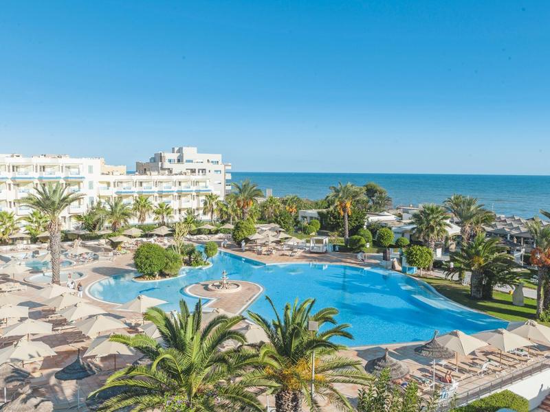 Grande piscina dell'hotel con lettini, palme e vista sul mare sotto un cielo limpido.