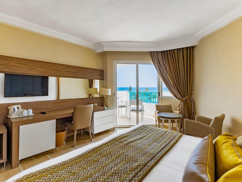 Camera d'hotel luminosa con vista mare, letto, scrivania, sedia e balcone con ringhiera in vetro.