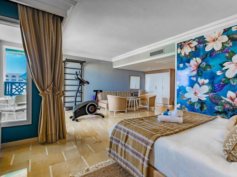 Camera d'hotel luminosa con letto matrimoniale, parete floreale, tavolo da pranzo e attrezzi da palestra.