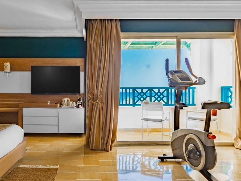 Camera d'hotel luminosa con letto, TV, area salotto e cyclette con vista sul mare.