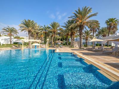 Großer Pool mit blauer Wasserfläche, umgeben von Palmen und Liegestühlen unter Sonnenschirmen.