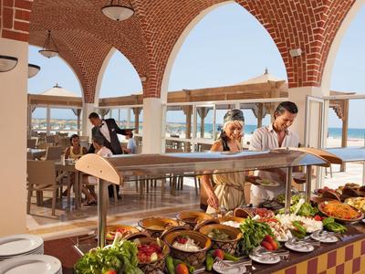 Buffet im Freien mit vielfältigen Speisen, Gäste nehmen sich Essen, mediterranes Ambiente am Meer.