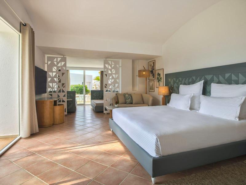 Ampia camera d'hotel con letto matrimoniale, divano, vista sul giardino, pavimento chiaro, arredamento moderno.