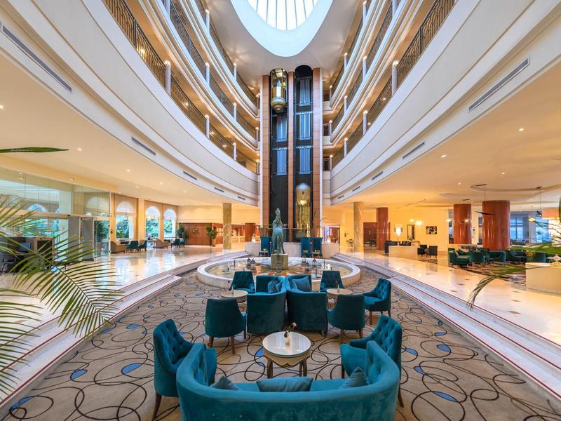 Atrio di hotel moderno con divani blu, ascensori centrali e luce naturale.