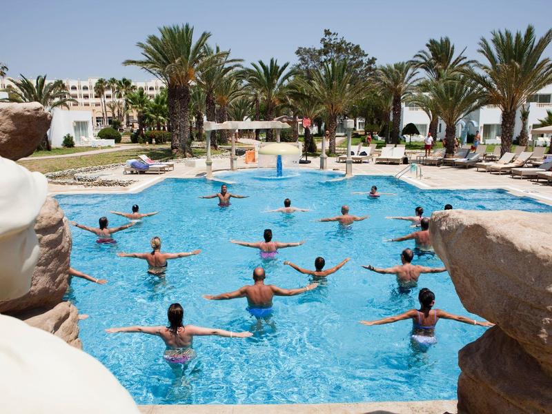 Persone che fanno acquagym in una piscina con palme e edifici bianchi intorno.