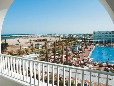 Vista da un balcone su una piscina dell'hotel, lettini e la spiaggia con il mare sullo sfondo.