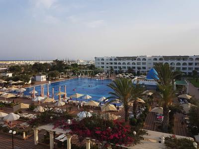 Grande piscina dell'hotel con ombrelloni, palme e piante in fiore sotto un cielo blu.
