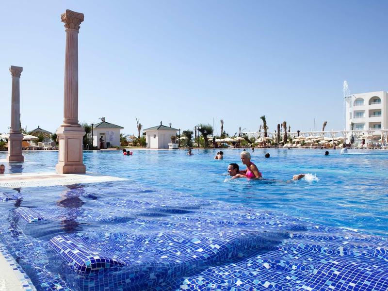 Piscina dell'hotel con colonne decorative e bagnanti sotto un cielo limpido.