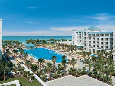 Luxury hotel with large pool, palm trees, and sea view under a clear blue sky.
