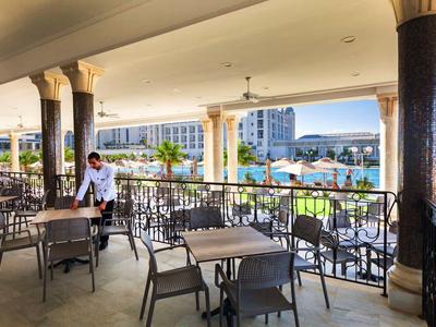 Offene Terrasse mit Metallmöbeln, Säulen und Blick auf Schwimmbad und Hotelgebäude im Hintergrund.