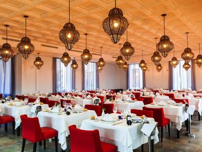 Großer, heller Speisesaal mit weißen Tischdecken, roten Stühlen und dekorativen hängenden Lampen.