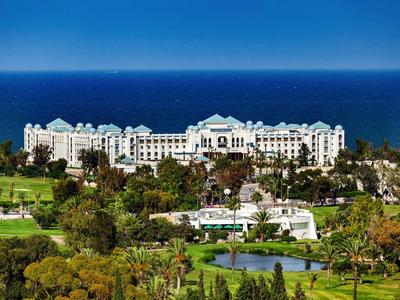 Großes weißes Hotel am Meer, umgeben von grünen Bäumen und einem Golfplatz mit Teich vorn.