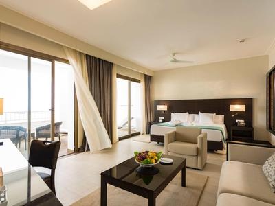 Bright hotel room with double bed, sofa, table, and sea view through large windows.