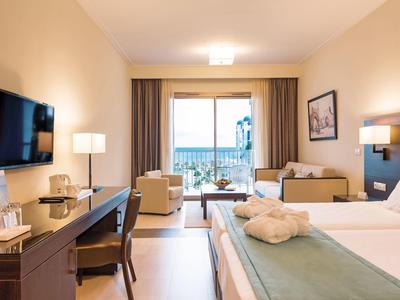 Modern hotel room with bed, desk, sofa, and balcony overlooking the sea.