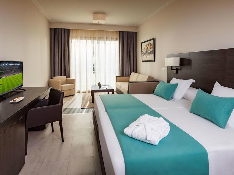 Modern hotel room with double bed, turquoise bedspread, and balcony with natural light.