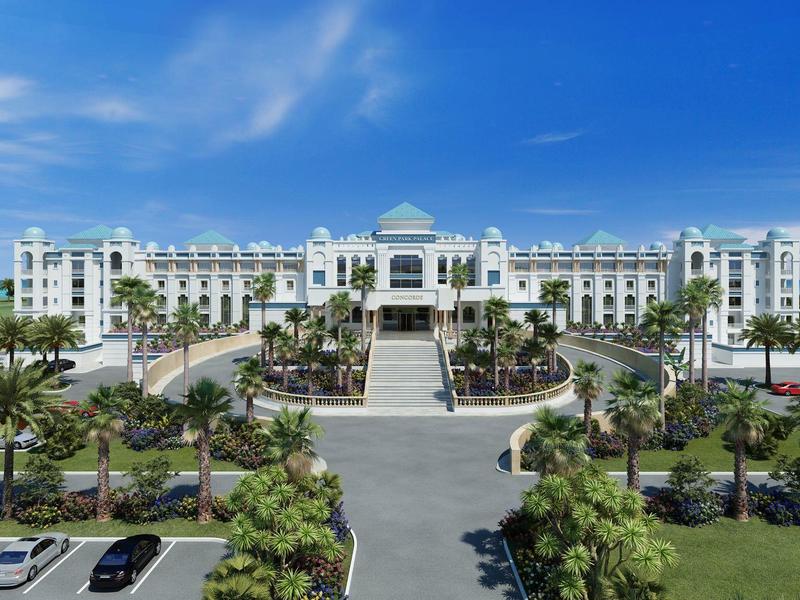Large white hotel building with palm trees and parking under a blue sky.