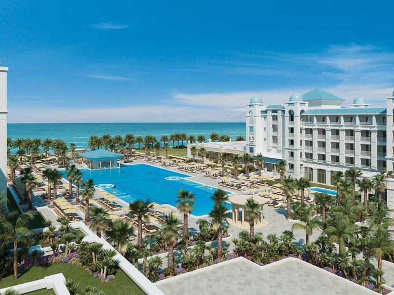 Luxury hotel with large pool, palm trees, and sea view under a clear blue sky.