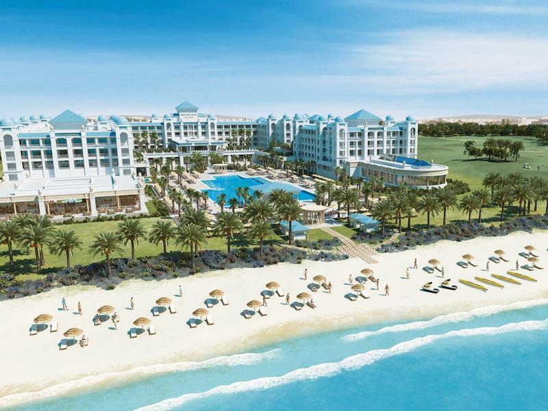 Luxury hotel with pool, palm trees, and sandy beach by blue sea under clear sky.