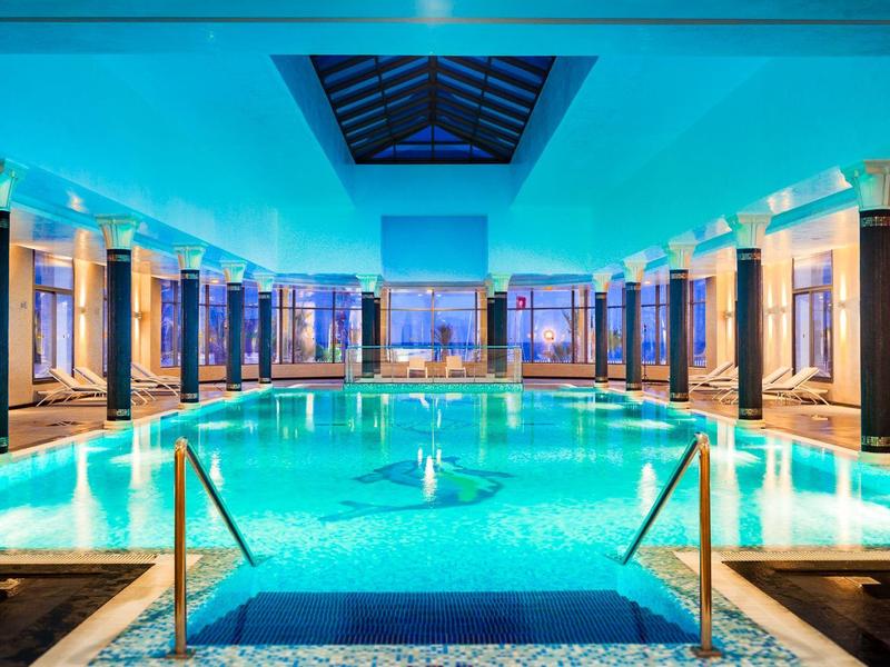 Modern indoor pool with blue-lit water and columns on the sides.