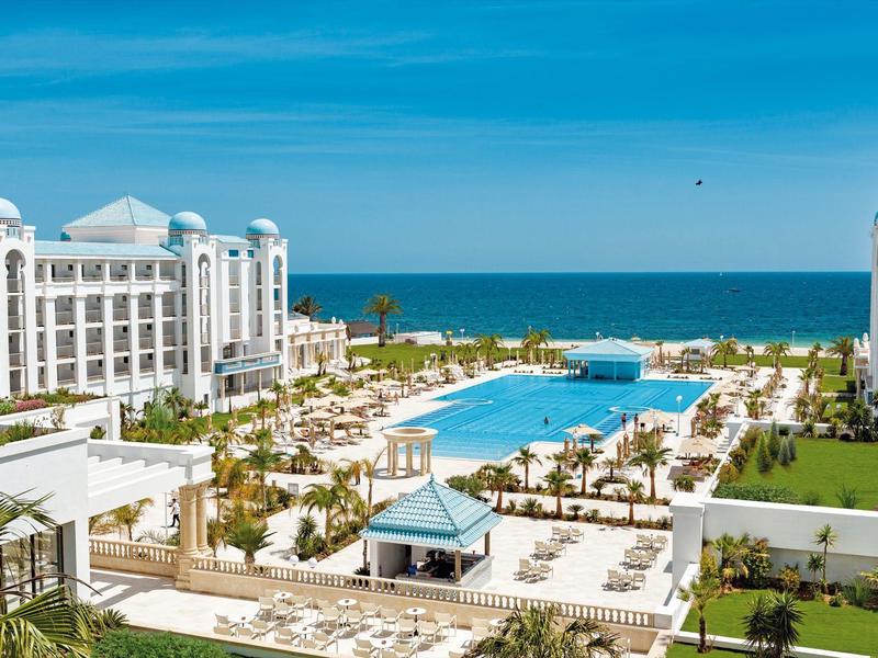 Large hotel resort with pool, sun umbrellas, and view of the blue sea under clear sky.